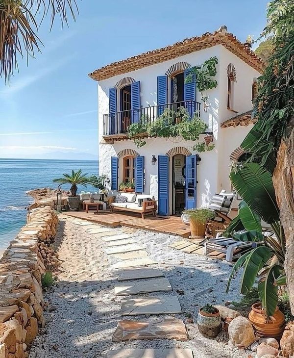 a white house with blue shutters next to the ocean and palm trees in front of it