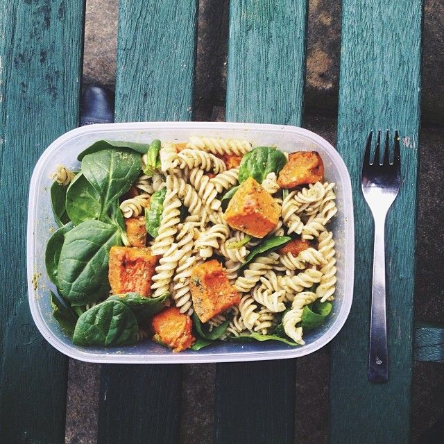 a plastic container filled with pasta and spinach on top of a wooden table next to a fork