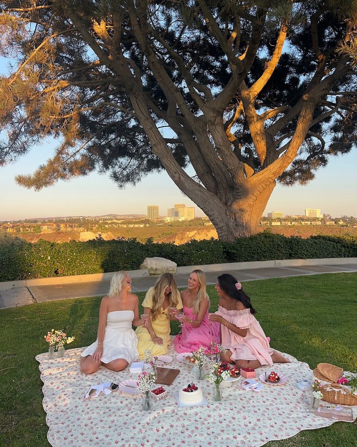 three women sitting on a blanket in front of a large tree eating cake and drinking tea