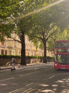 a red double decker bus driving down a street next to tall buildings with trees on both sides