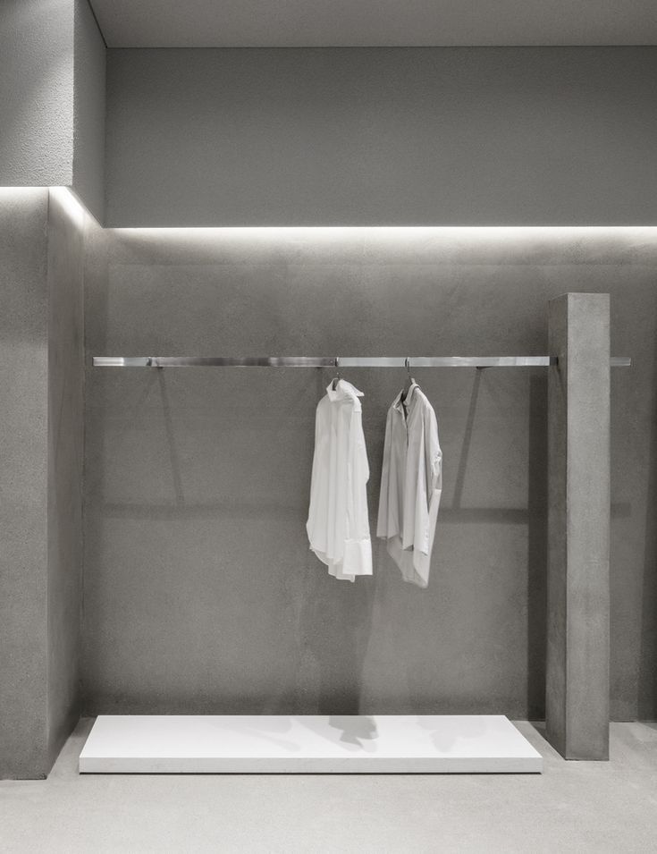 two white shirts are hanging on a clothes rack in a room with concrete walls and flooring