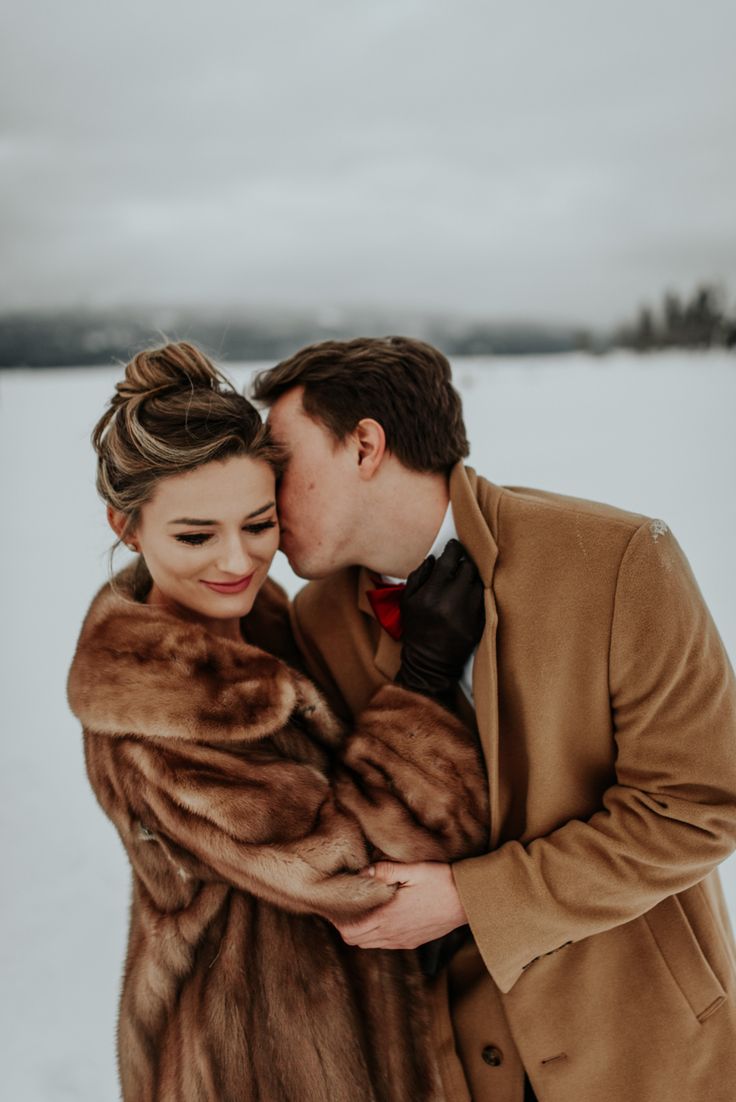 a man and woman are standing in the snow hugging each other while wearing fur coats