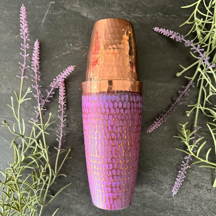 a pink and gold vase sitting next to some purple flowers on a gray surface with leaves