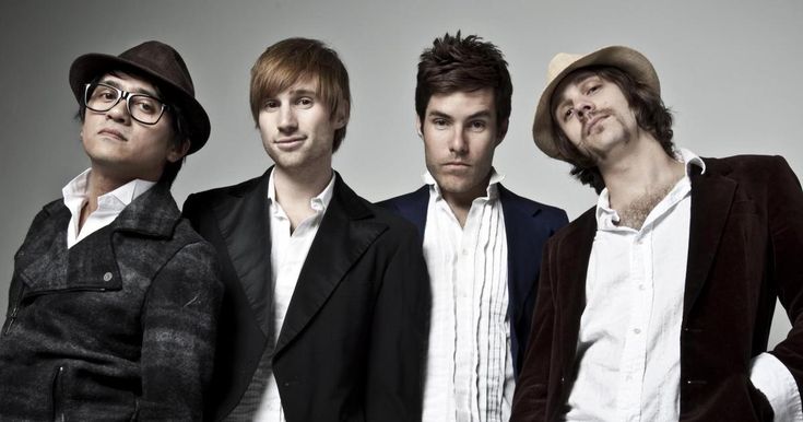 four young men are posing for a photo together, one is wearing a fedora and the other wears a blazer