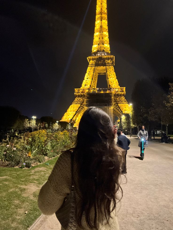 a woman standing in front of the eiffel tower at night with her back to the camera