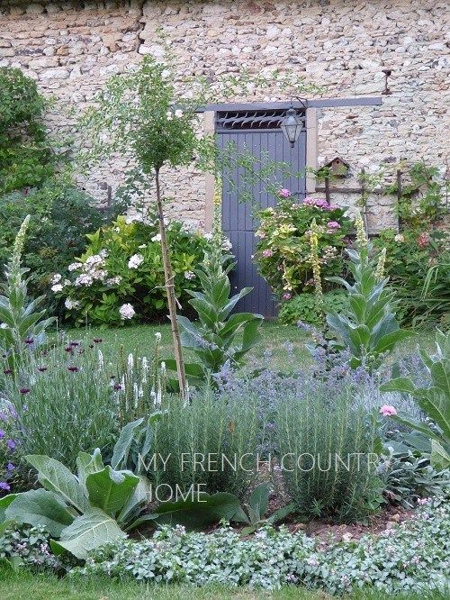 a garden with lots of flowers and plants around it in front of a stone building