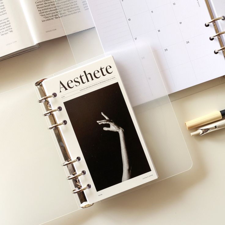 an open notebook with a hand on it next to a pen and calendars that read aesthete