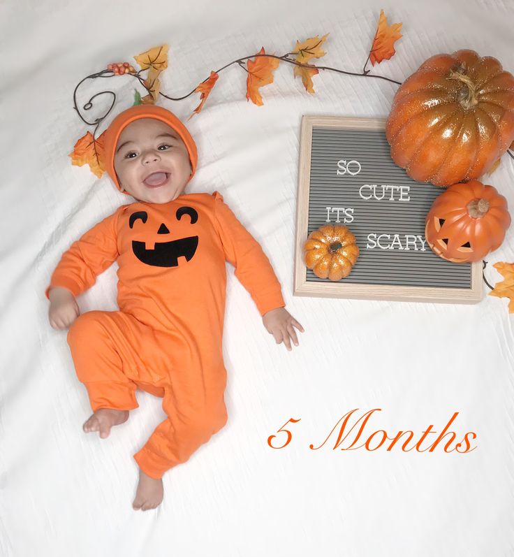 a baby in an orange pumpkin costume laying on a bed next to some pumpkins