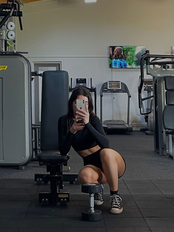 a woman taking a selfie with her phone in a gym area while sitting on a bench