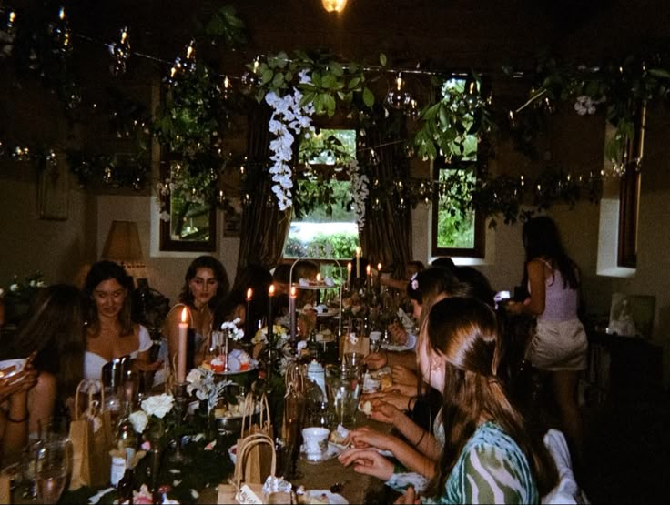 a group of people sitting around a long table with candles on it and flowers hanging from the ceiling