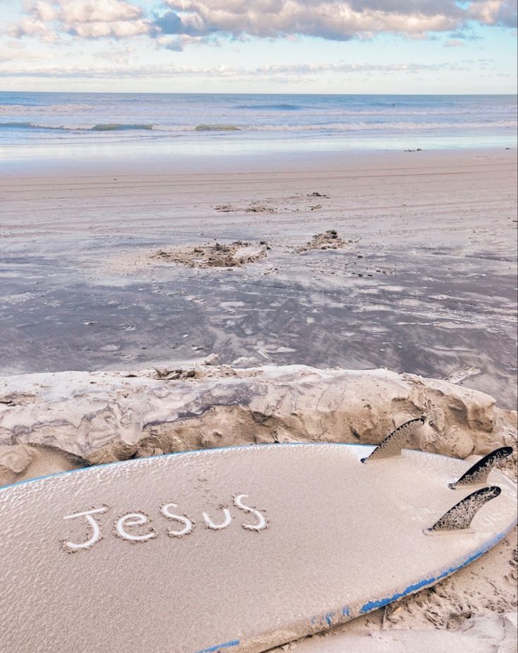 a surfboard that is laying in the sand with jesus written on it and water behind it