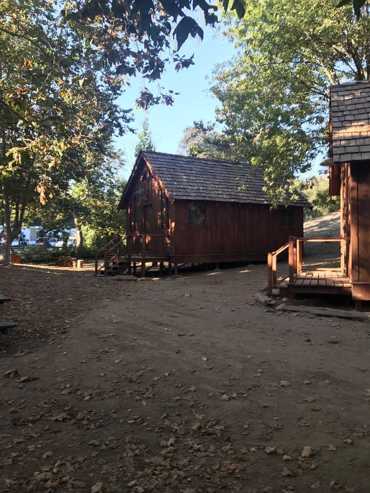 two small wooden buildings sitting on top of a dirt field next to trees and benches