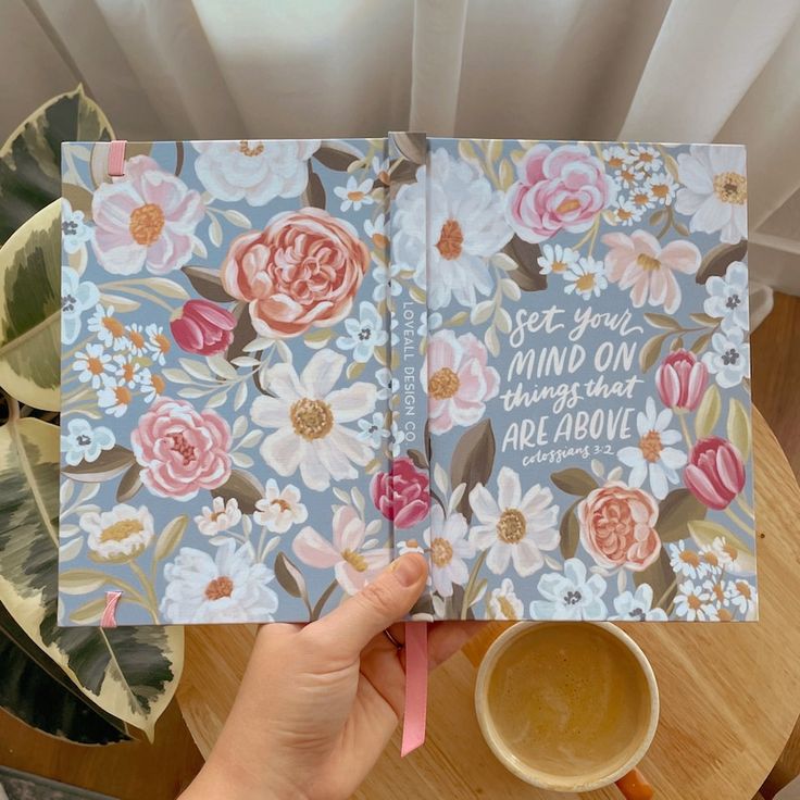 a person holding up a book with flowers on it and a cup of coffee in front of them