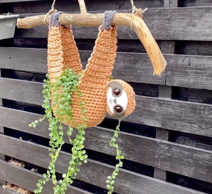 a crocheted sloth hanging from a tree branch with green leaves on it