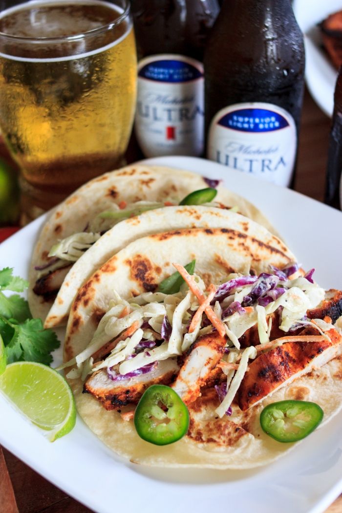 three tacos on a white plate with limes and beer in the background,