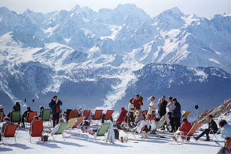 a group of people sitting in lawn chairs on top of a snow covered slope with mountains in the background