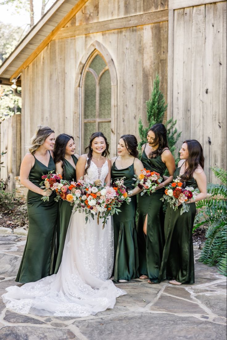 a group of women standing next to each other in front of a wooden building holding bouquets