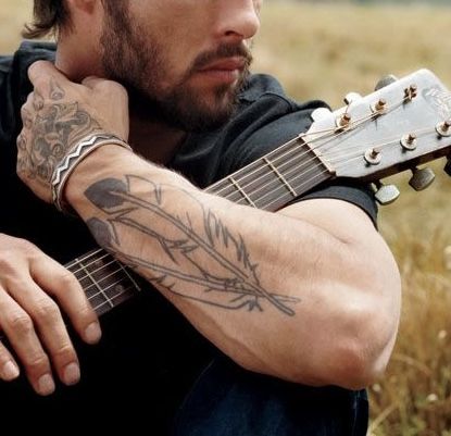 a man with tattoos on his arm holding an acoustic guitar in the grass and looking off into the distance