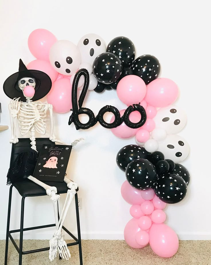 a skeleton sitting on a chair in front of a balloon wall with the word boo spelled out