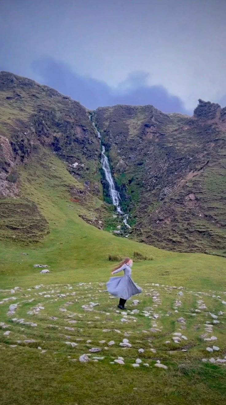 a woman standing on top of a lush green hillside next to a waterfall holding an umbrella