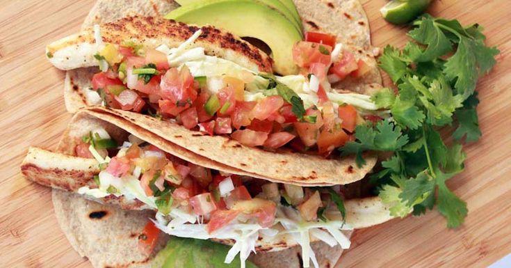 three tacos on a wooden cutting board with avocado and salsa in the middle