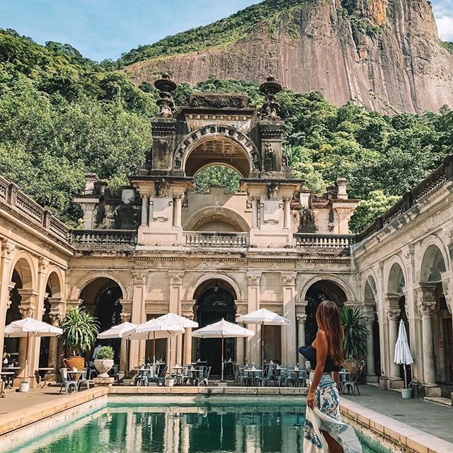 a woman standing in front of a pool surrounded by mountains and greenery with umbrellas