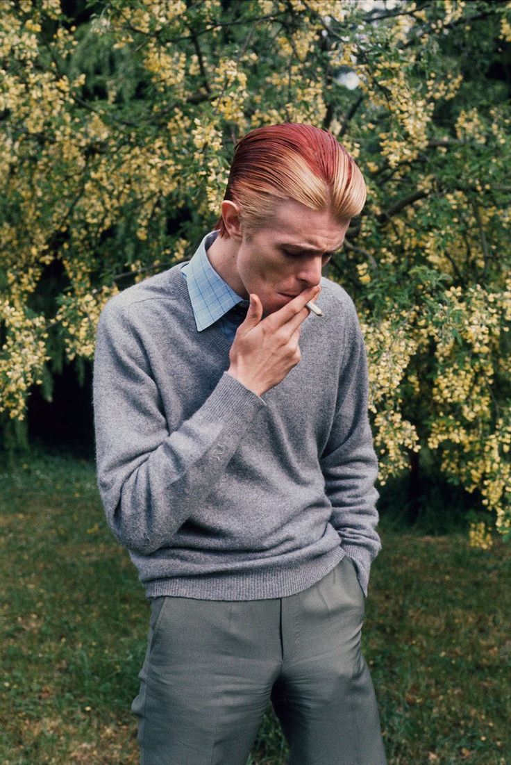 a man with red hair is standing in front of a tree and holding his hand to his face