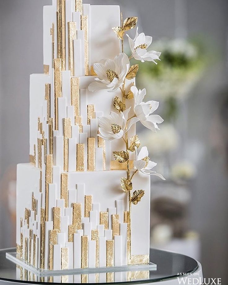 a white and gold wedding cake with flowers on the top is sitting on a glass table