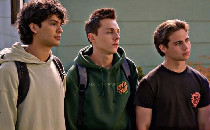 three young men standing next to each other in front of a building and one is wearing a backpack