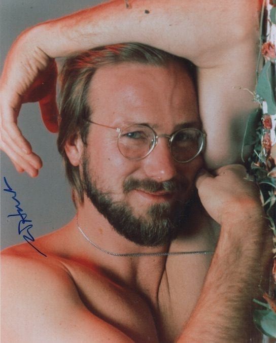 a shirtless man with glasses leaning against a wall and holding his hand on his head