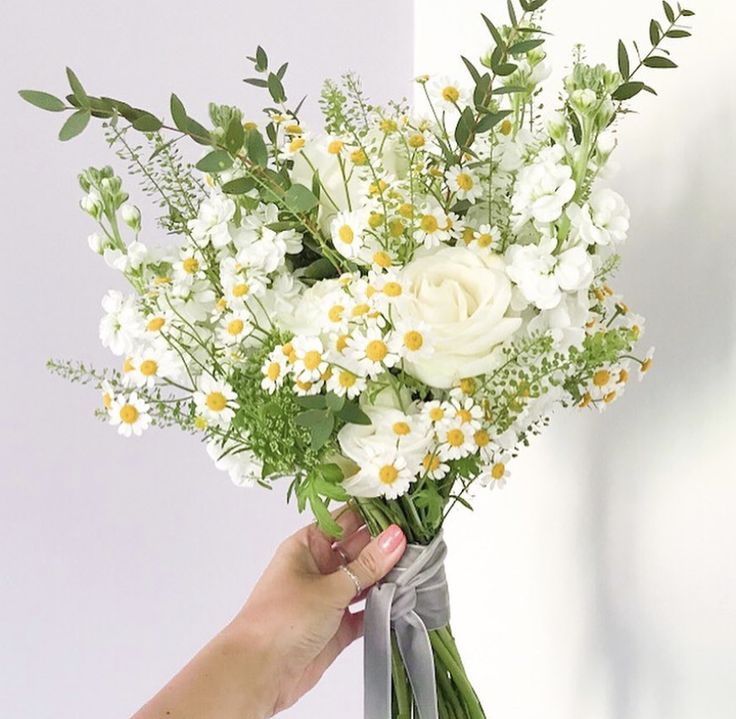 a woman's hand holding a bouquet of white and yellow flowers with greenery