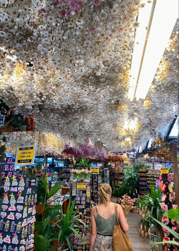 a woman walking through a store filled with lots of plants and hanging flowers on the ceiling