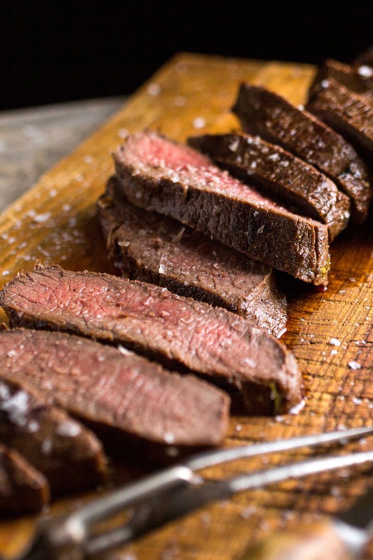 sliced steak sitting on top of a wooden cutting board next to a knife and fork