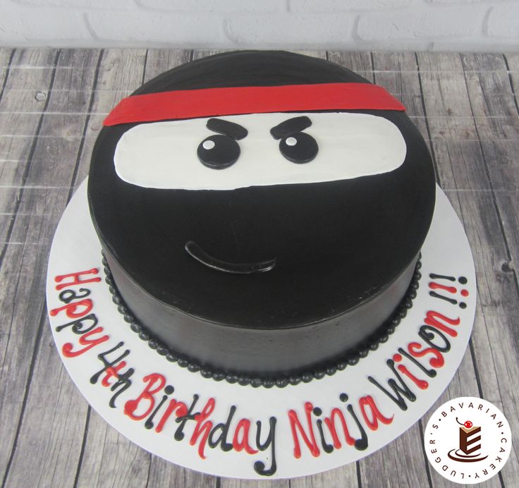 a black and white birthday cake with a ninja face on it's top is sitting on a wooden table