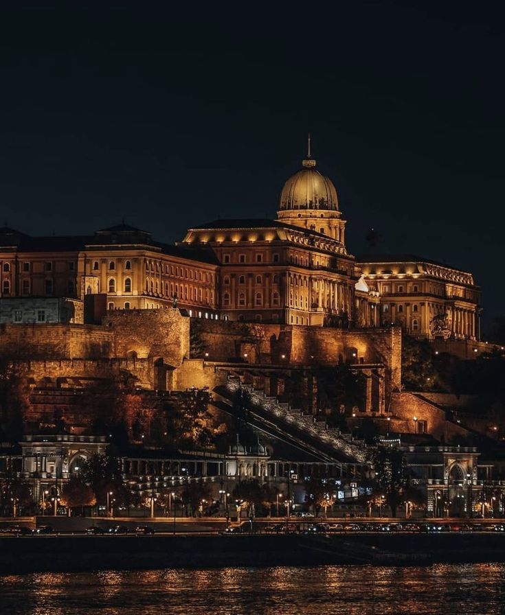 a large building on top of a hill next to the water at night with lights on it