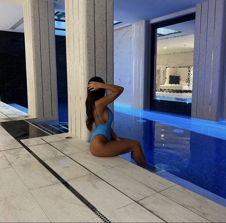 a beautiful woman in a blue swimsuit sitting next to a swimming pool with columns