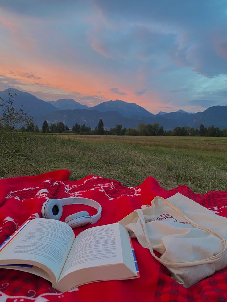 an open book sitting on top of a red blanket next to a bag and headphones