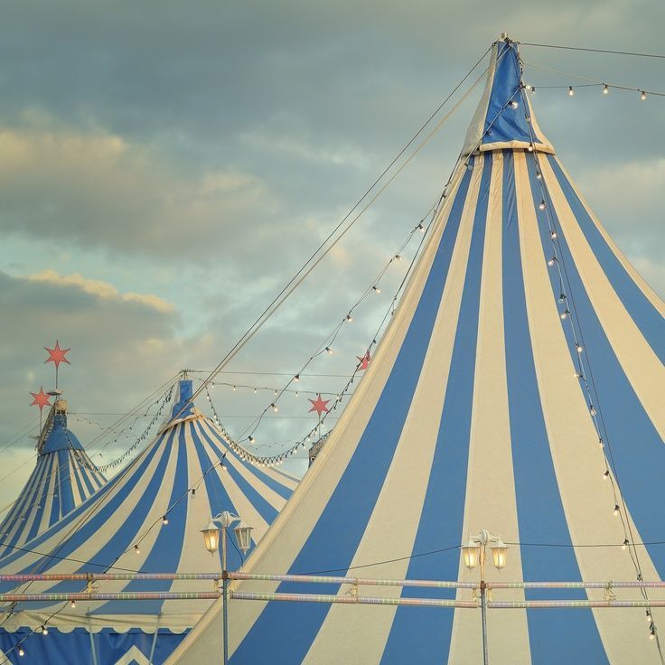 a large blue and white circus tent with string lights on it's sides under cloudy skies