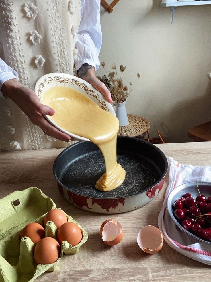 a person pouring batter into a pan with cherries on the table next to it