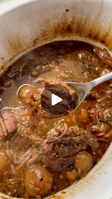 a pot filled with stew and meat on top of a wooden table next to a spoon