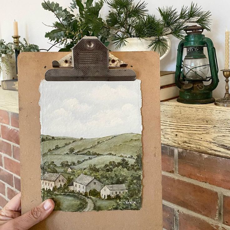 a hand holding a clipboard with a painting on it next to a brick wall