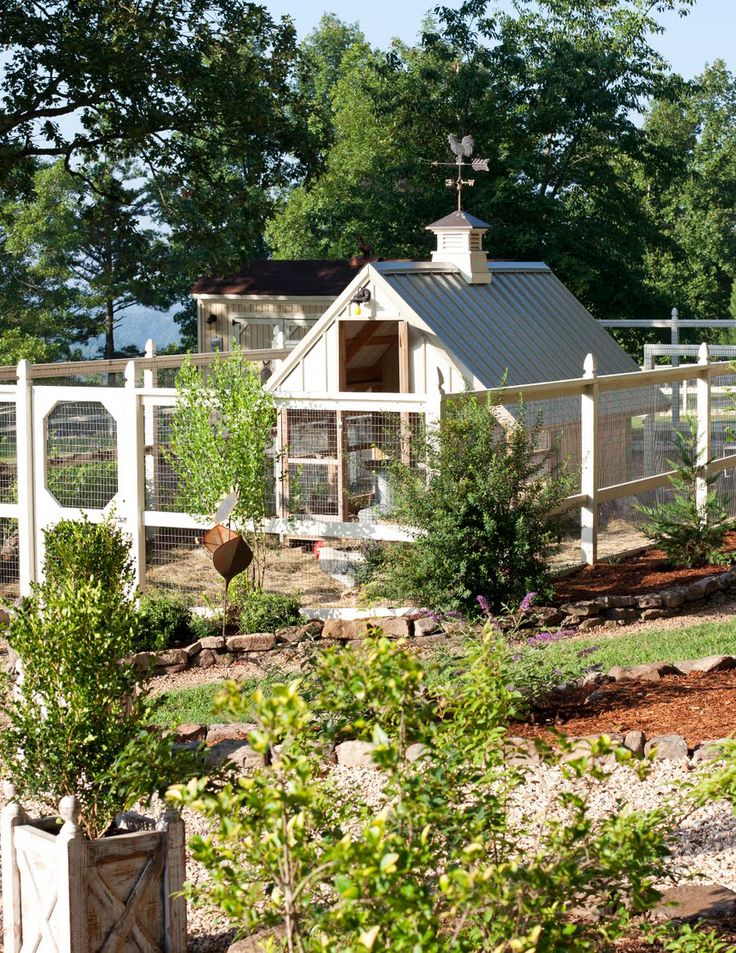 a chicken coop in the middle of a garden with trees and shrubs around it, next to a white picket fence