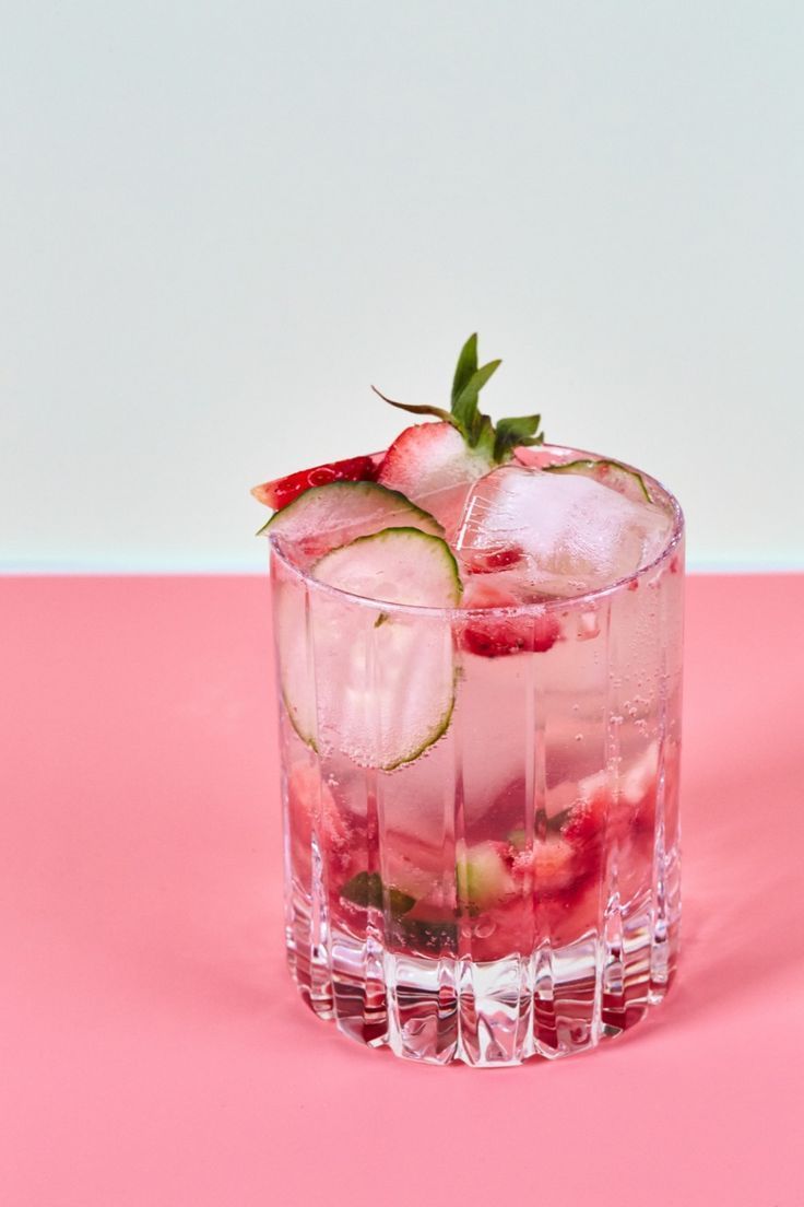 a glass filled with ice and strawberries on top of a pink surface