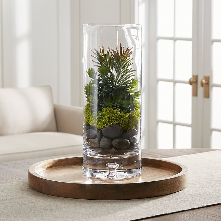 a glass vase filled with plants and rocks on top of a wooden tray next to a couch