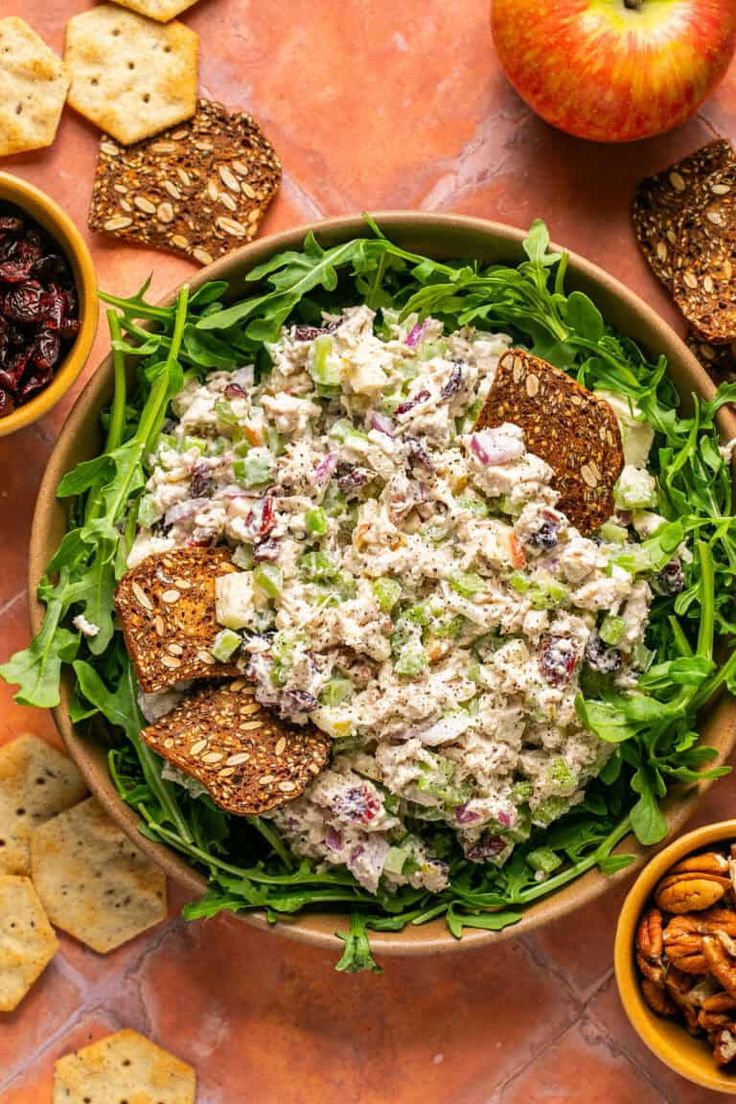 a bowl filled with salad surrounded by crackers and fruit on the side, next to an apple