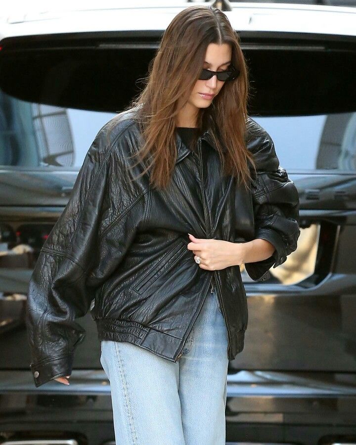 a woman in black leather jacket and jeans walking towards a car with her hand on her hip