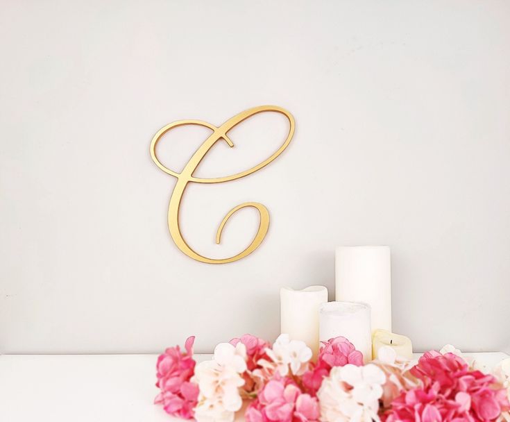 flowers and candles sit on a table with a gold monogrammed wall hanging in the background