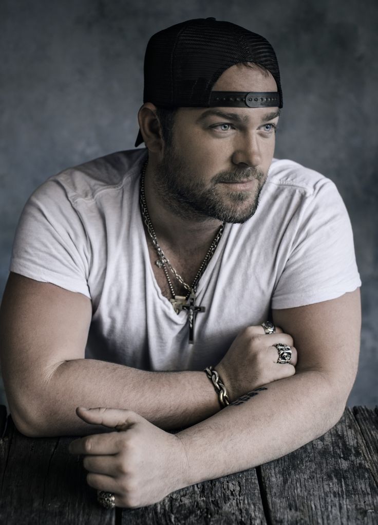 a man with his arms crossed sitting on a wooden table wearing a white shirt and black hat