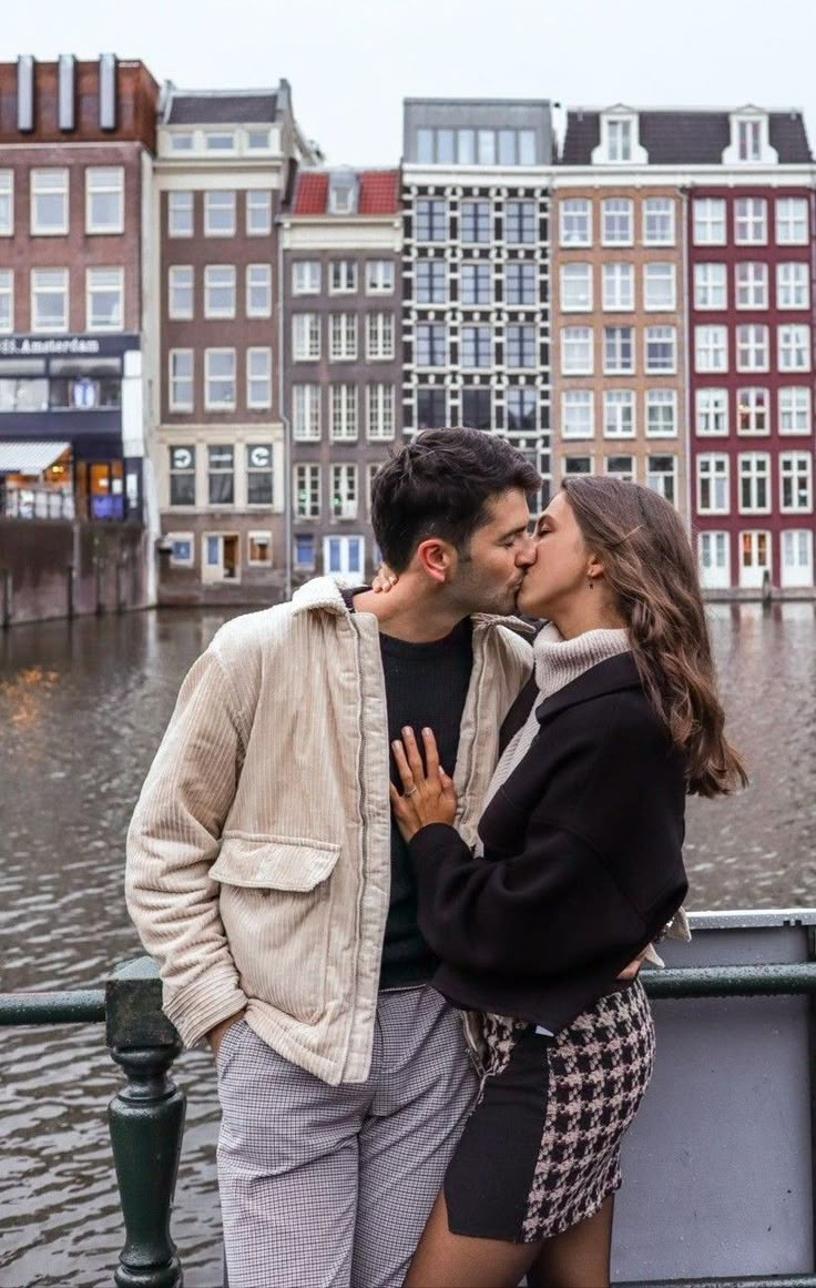 a man and woman standing next to each other in front of water with buildings behind them