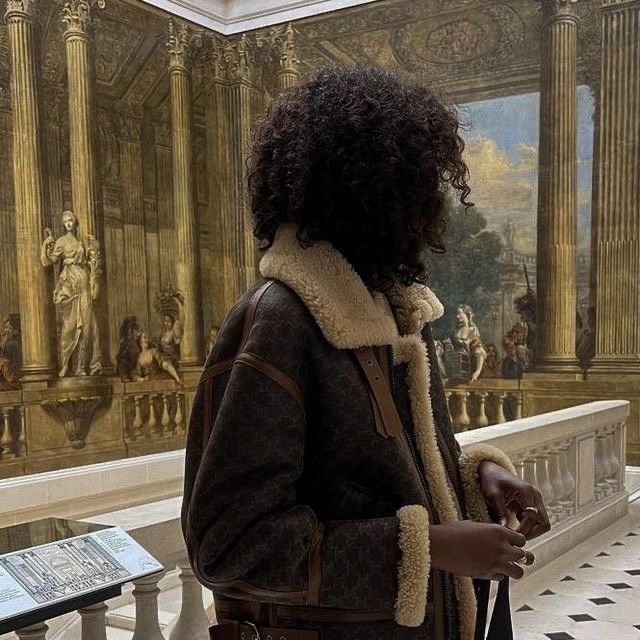 a woman is standing in front of a painting and looking at her cell phone while wearing a brown jacket